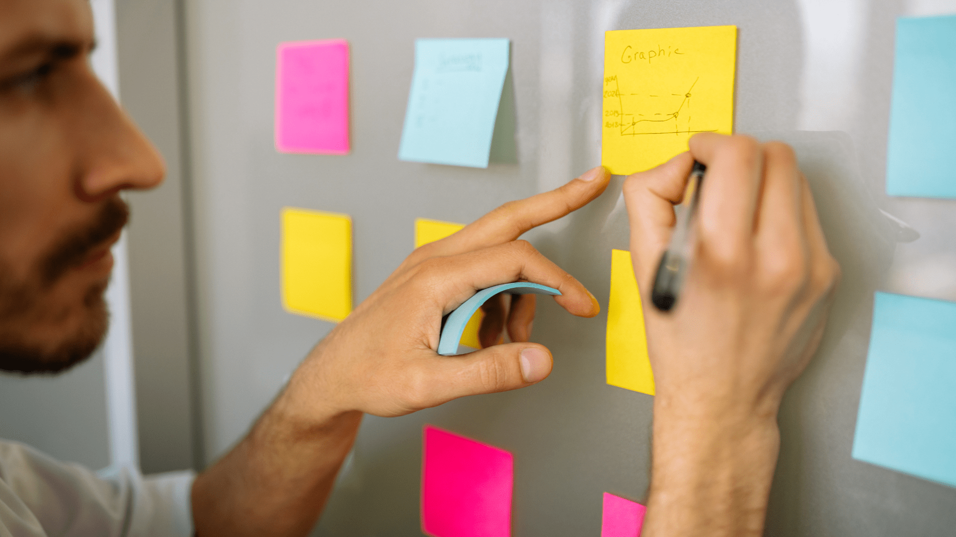 Eine Person schreibt mit einem Stift auf ein Post-It an der Wand